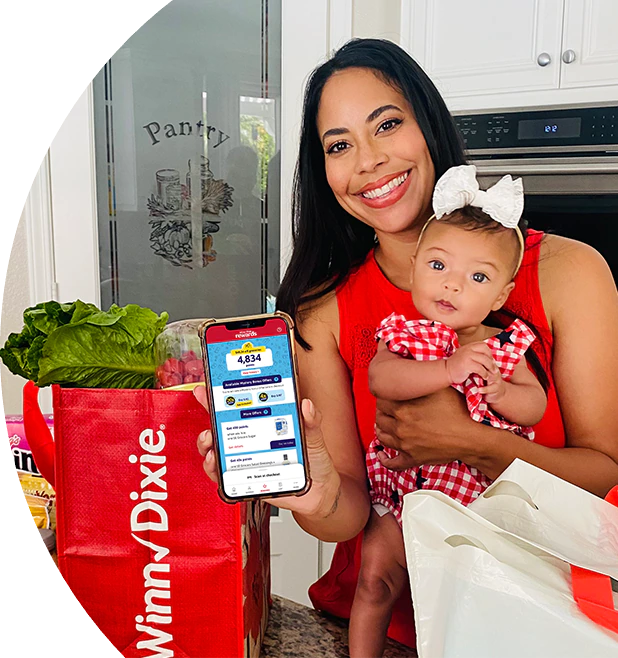 Woman in kitchen holding a baby and displaying her phone with the Winn-Dixie app.