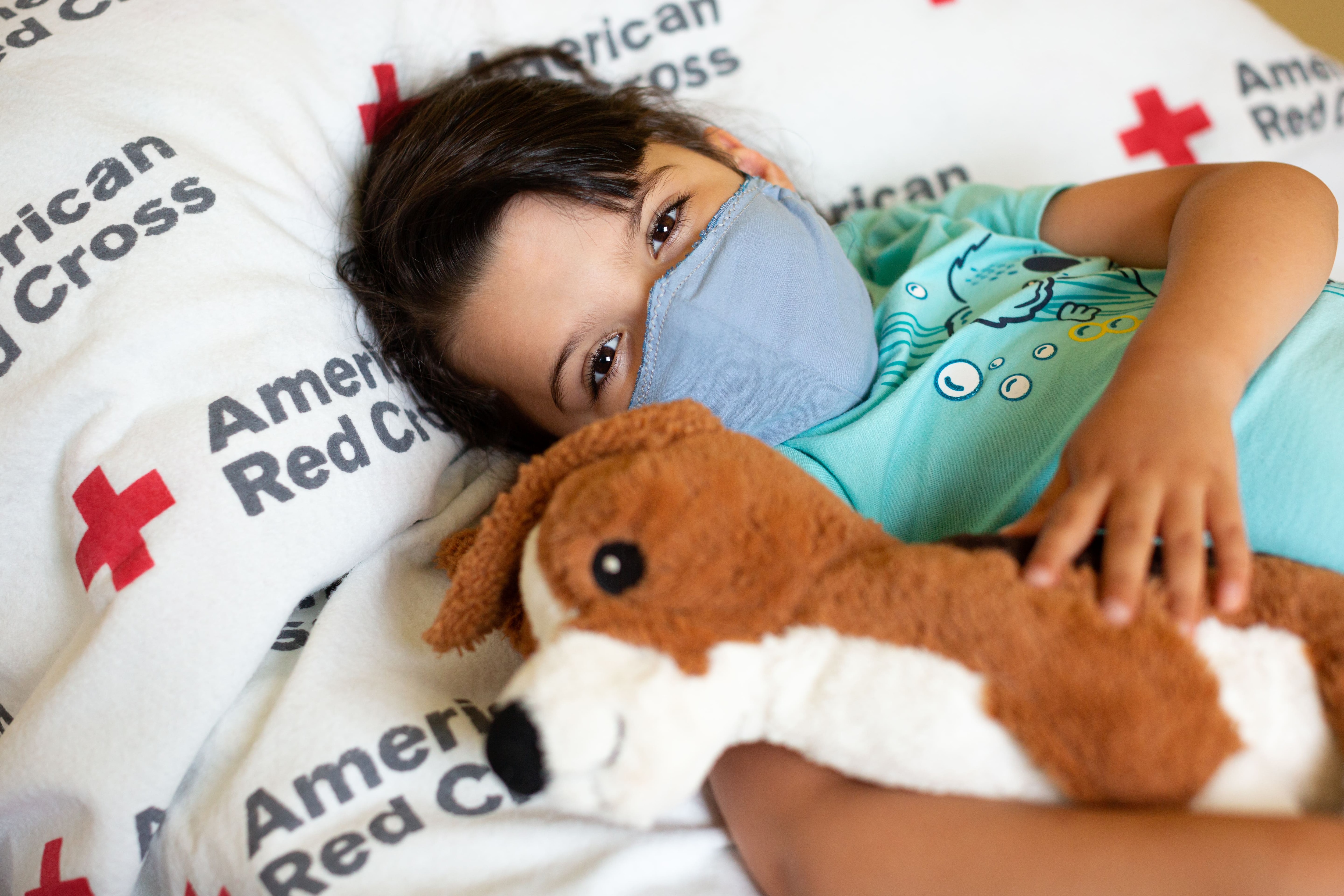 Little kid wearing a mask on American Red Cross bed with a stuffed animal