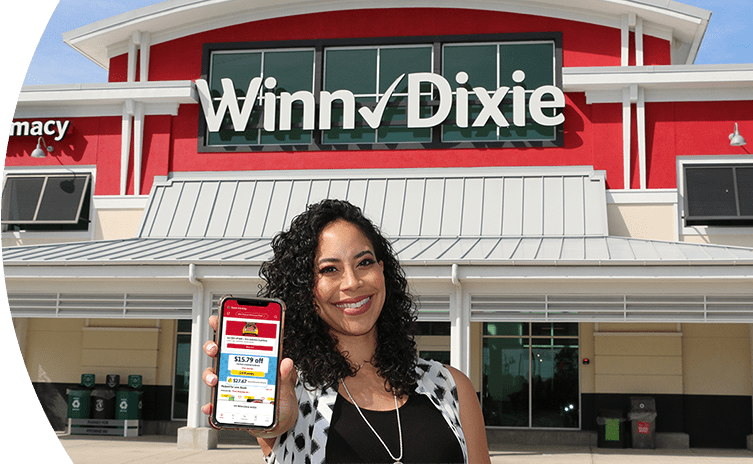 Smiling woman standing in front of a Winn-Dixie. She is holding up her phone with the screen showing the Winn-Dixie app.