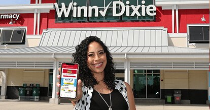 Smiling woman standing in front of a Winn-Dixie. She is holding up her phone with the screen showing the Winn-Dixie app.
