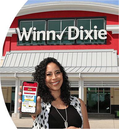 Smiling woman standing in front of a Winn-Dixie. She is holding up her phone with the screen showing the Winn-Dixie app.