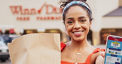 Woman smiling outside of a Winn-Dixie holding a grocery bag and phone with Winn-Dixie app