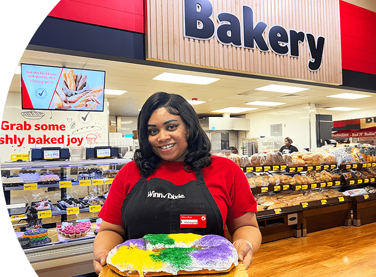Winn-Dixie associate standing in front of the in-store bakery. She is smiling and holding a Mardi Gras cake.