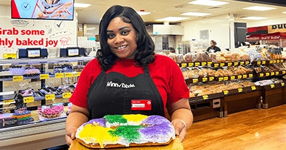 Winn-Dixie associate standing in front of the in-store bakery. She is smiling and holding a Mardi Gras cake.