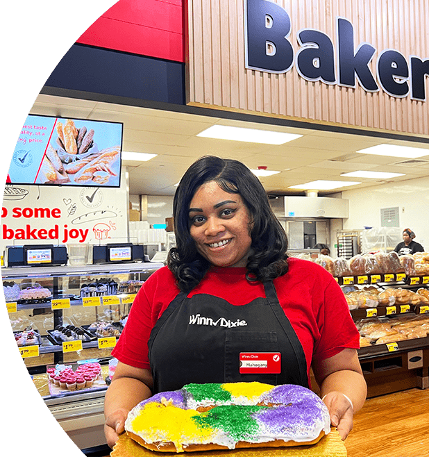 Winn-Dixie associate standing in front of the in-store bakery. She is smiling and holding a Mardi Gras cake.
