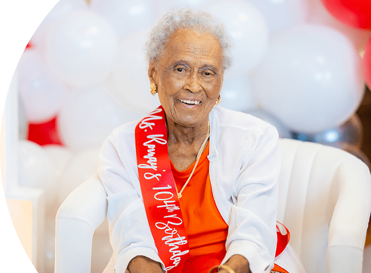 Romay Davis sitting and smiling. She is wearing a ribbon that says "Ms. Romay's 104th Birthday".