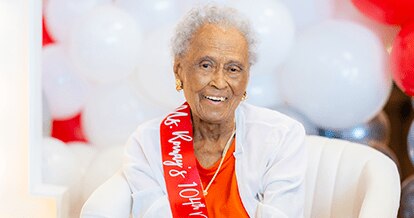Romay Davis sitting and smiling. She is wearing a ribbon that says "Ms. Romay's 104th Birthday".