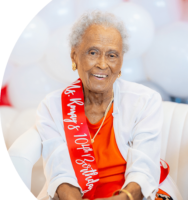 Romay Davis sitting and smiling. She is wearing a ribbon that says "Ms. Romay's 104th Birthday".