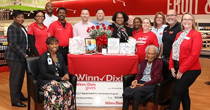 Mrs. Romay Davis joyfully sitting on a chair smiling and surrounded by Winn-Dixie gives foundation members.