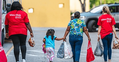 Two Winn-Dixie volunteers with a mom and a daughter carrying groceries