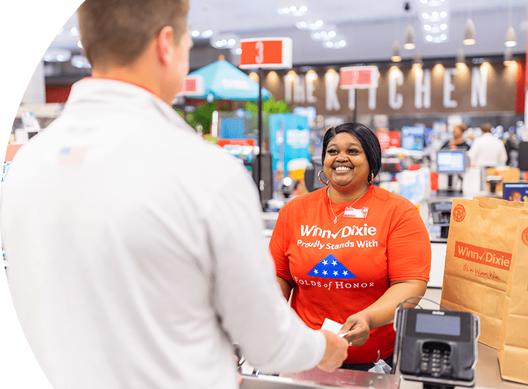 Smiling Winn-Dixie cashier wearing a Folds of Honor shirt. On the foreground, a customer is making a transaction.