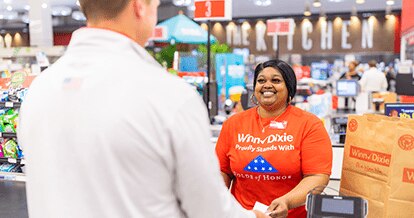 Smiling Winn-Dixie cashier wearing a Folds of Honor shirt. On the foreground, a customer is making a transaction.