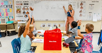 Teacher in the front of class pointing at a whiteboard. In front of her, young students are sitting on desks and raising their hands. On one of the desks there is a red Winn-Dixie reusable bag.