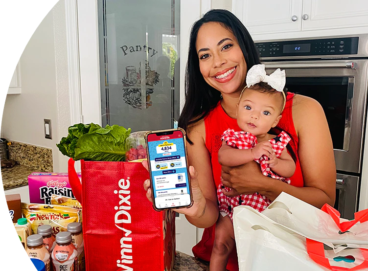 Woman in kitchen holding a baby and displaying her phone with the Winn-Dixie app.