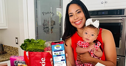 Woman in kitchen holding a baby and displaying her phone with the Winn-Dixie app.