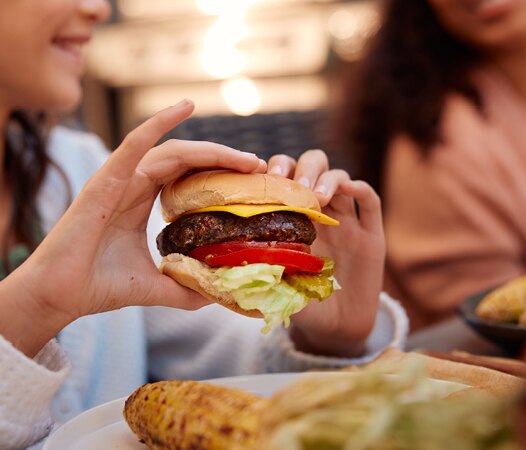 A person holding a cheeseburger with lettuce, tomato, and a beef patty in a soft bun. An outdoor meal, with another person blurred in the background and grilled corn and other food visible on the table.