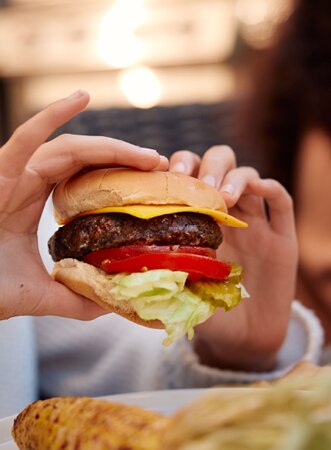 A person holding a cheeseburger with lettuce, tomato, and a beef patty in a soft bun. An outdoor meal, with another person blurred in the background and grilled corn and other food visible on the table.