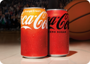 Two Coca-Cola cans placed on a basketball court, with a crowd and a large basketball visible in the blurred background. The can on the left is Coca-Cola Orange Cream, with a gradient from orange to red and white branding. The can on the right is Coca-Cola Zero Sugar, in a solid red color with black branding.