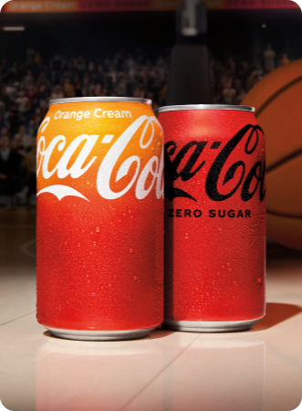 Two Coca-Cola cans placed on a basketball court, with a crowd and a large basketball visible in the blurred background. The can on the left is Coca-Cola Orange Cream, with a gradient from orange to red and white branding. The can on the right is Coca-Cola Zero Sugar, in a solid red color with black branding.