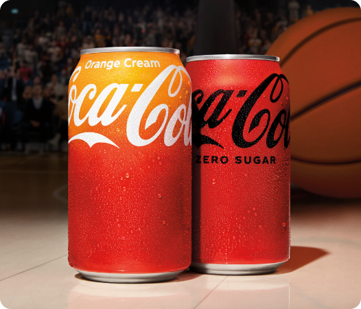 Two Coca-Cola cans placed on a basketball court, with a crowd and a large basketball visible in the blurred background. The can on the left is Coca-Cola Orange Cream, with a gradient from orange to red and white branding. The can on the right is Coca-Cola Zero Sugar, in a solid red color with black branding.