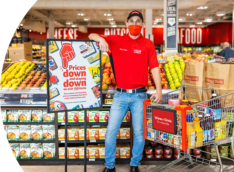 Happy Winn-Dixie employee standing in-between a shopping cart, and a "Prices down and staying down" sign