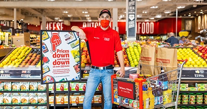 Happy Winn-Dixie employee standing in-between a shopping cart, and a "Prices down and staying down" sign