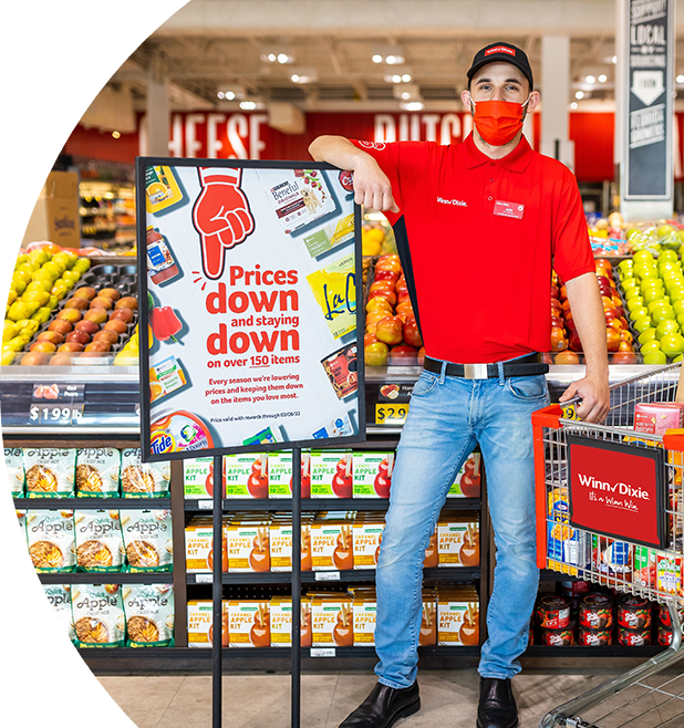 Happy Winn-Dixie employee standing in-between a shopping cart, and a "Prices down and staying down" sign